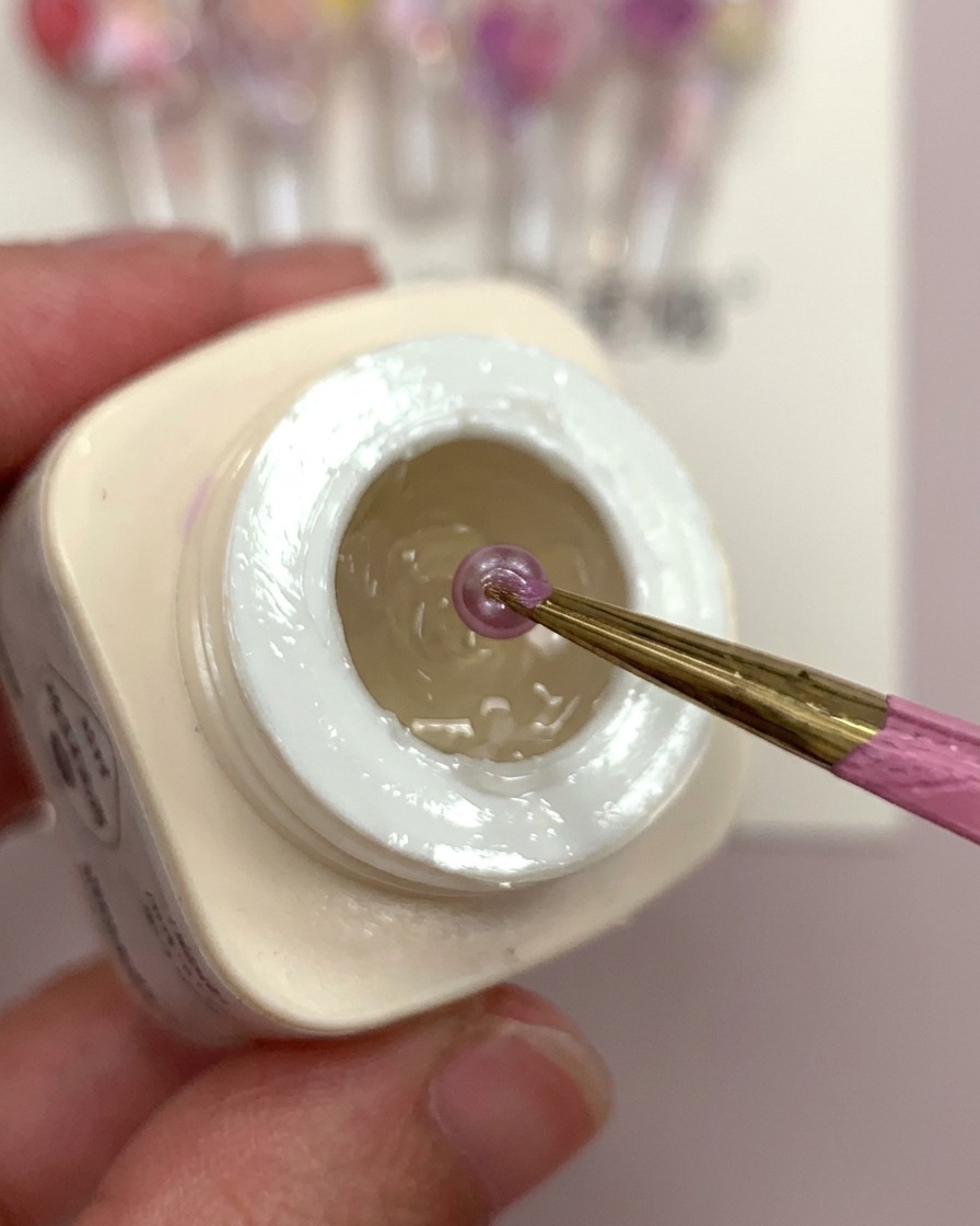 Eine Hand hält eine klare Nagelspitze mit einem Tropfen Frosted-Gel, in den lila Blumenkugeln eingesetzt werden.