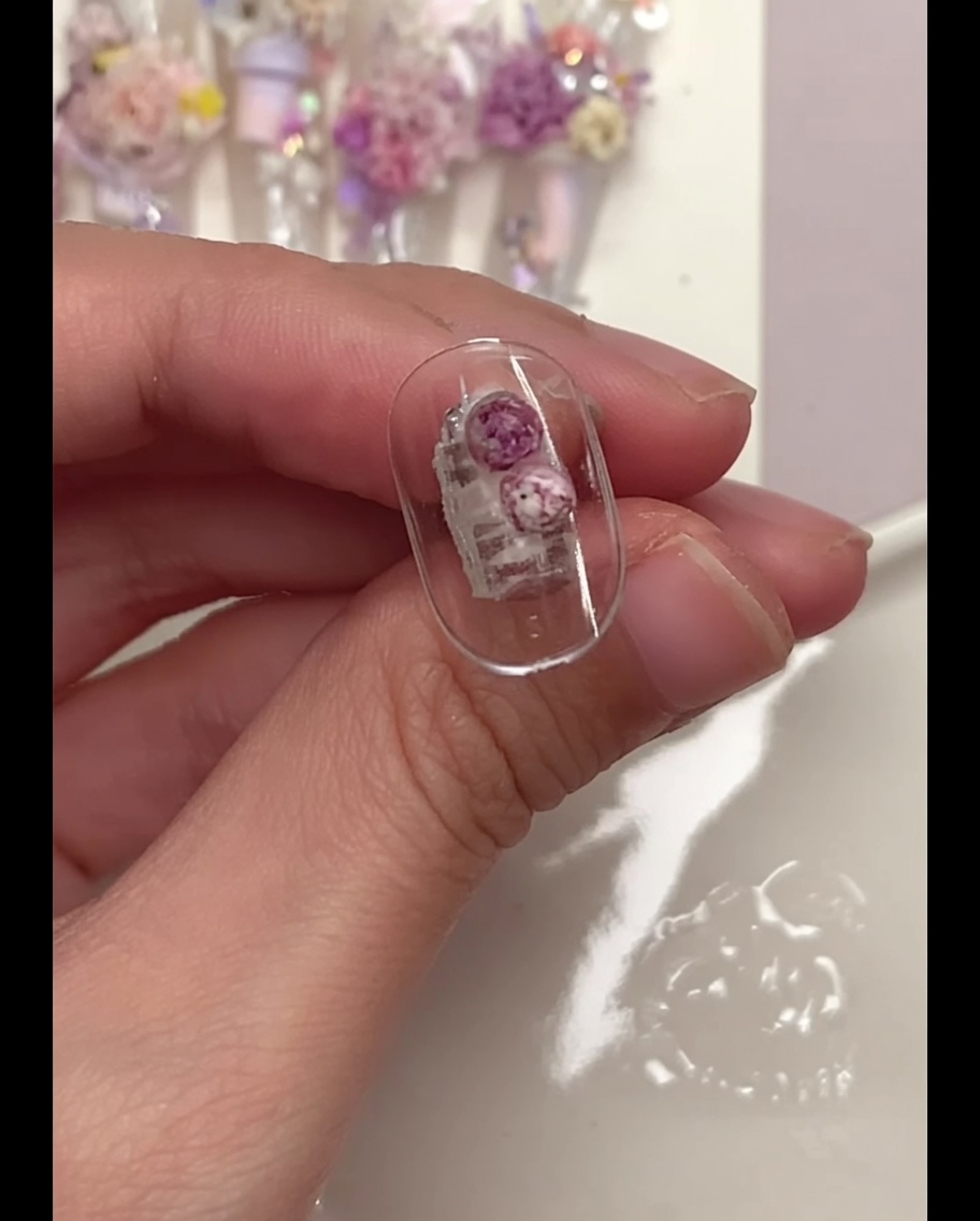 Ein Detailpinsel drückt vorsichtig auf lila Blumenkugeln, die auf einer klaren Nagelspitze platziert wurden.