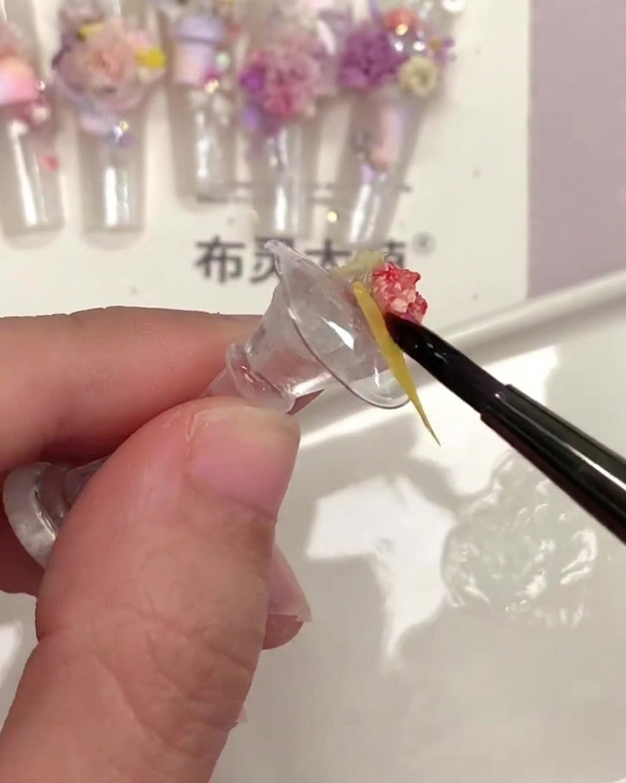 Ein feiner Nagelpinsel trägt klaren Top-Coat unter ein kleines gelbes Trockenblumenstück auf.