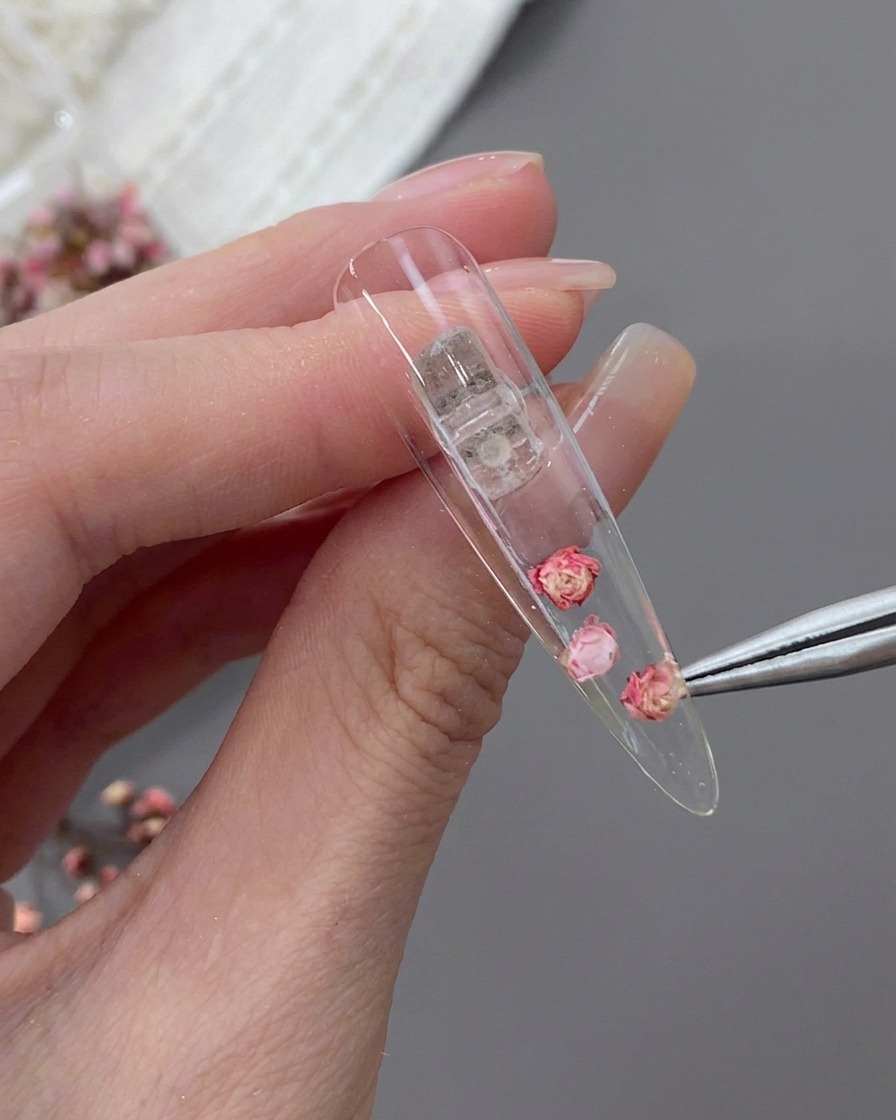 Eine Pinzette platziert kleine rosa und weiße Trockenblumen um die zentrale Rose auf dem Nageltip.