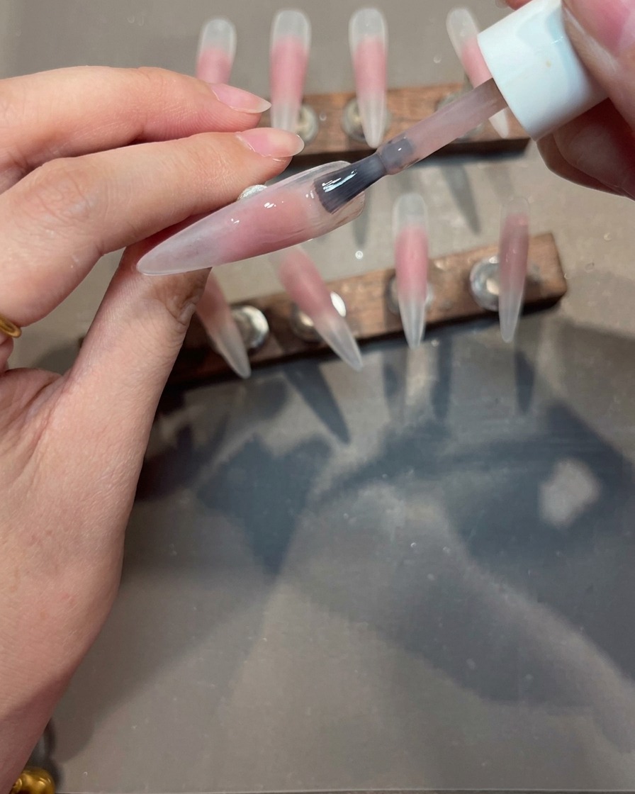 Ein Pinsel trägt eine glatte Schicht Nude-Gel auf einen Nageltipp mit einem rosa Blush-Zentrum auf.