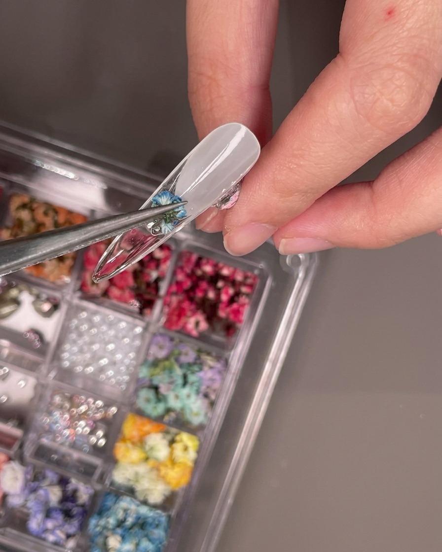 Eine Pinzette platziert eine kleine blaue Trockenblume auf der Unterseite eines klaren Nageltips.