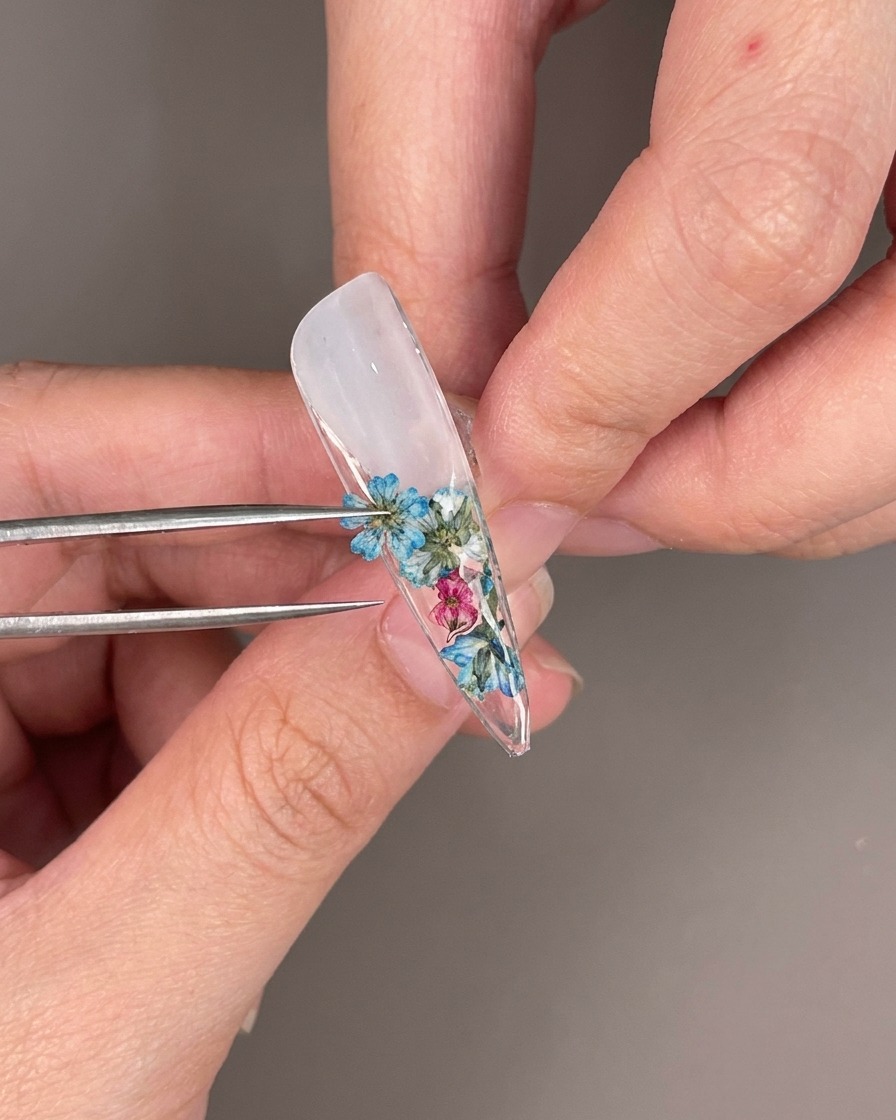 Nahaufnahme von klaren Strasssteinen, die zwischen Trockenblumen auf der Unterseite eines Nageltips platziert werden.