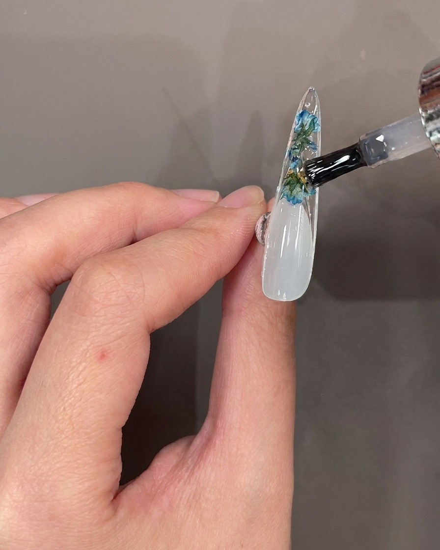 Ein Nagelpinsel trägt klaren Top Coat über Blüten und Steine auf der Unterseite eines Nageltips auf.