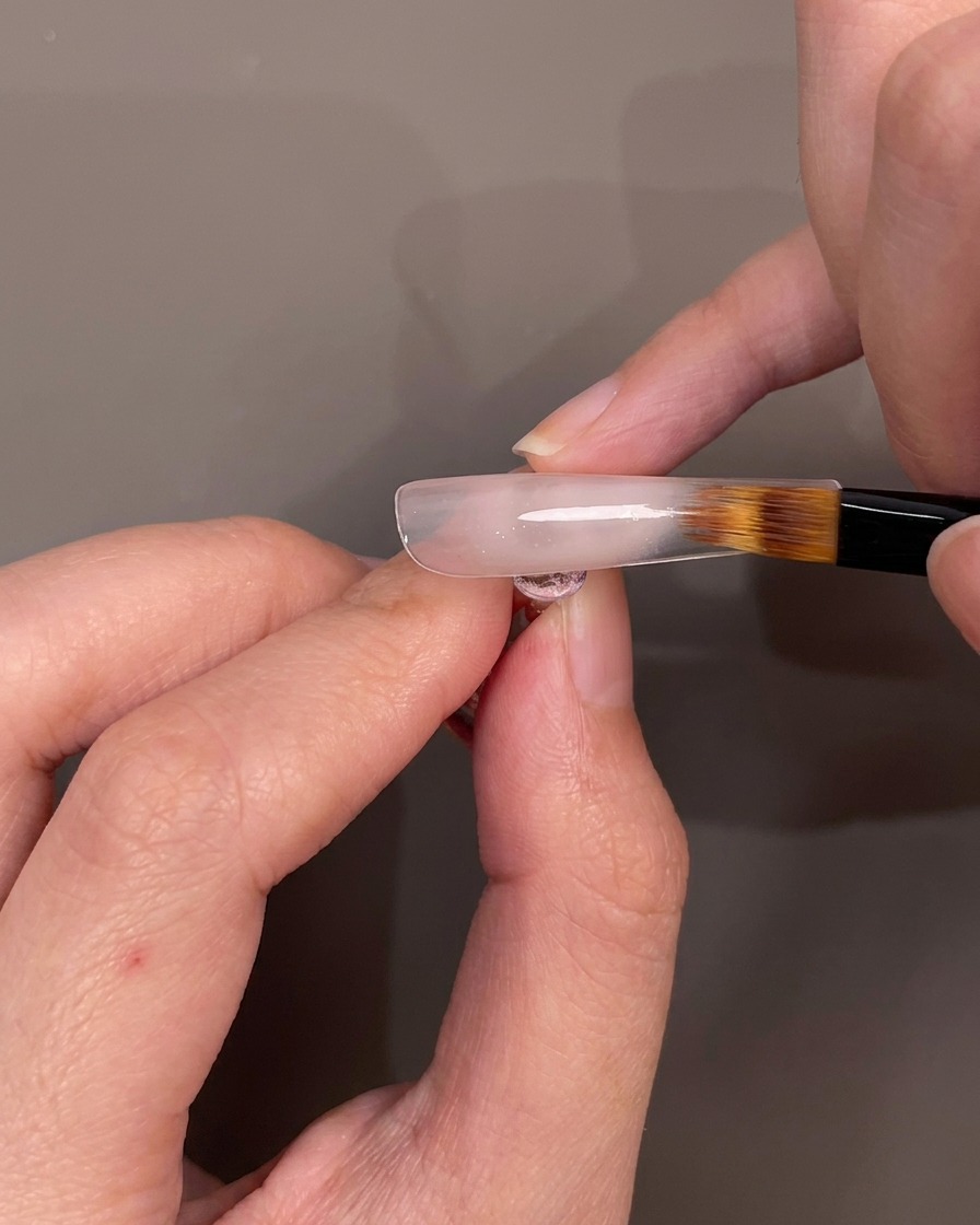 Ein Pinsel verblendet nudefarbenes Gel auf einem Nageltip nach unten, um einen Ombré-Effekt zu erzielen.