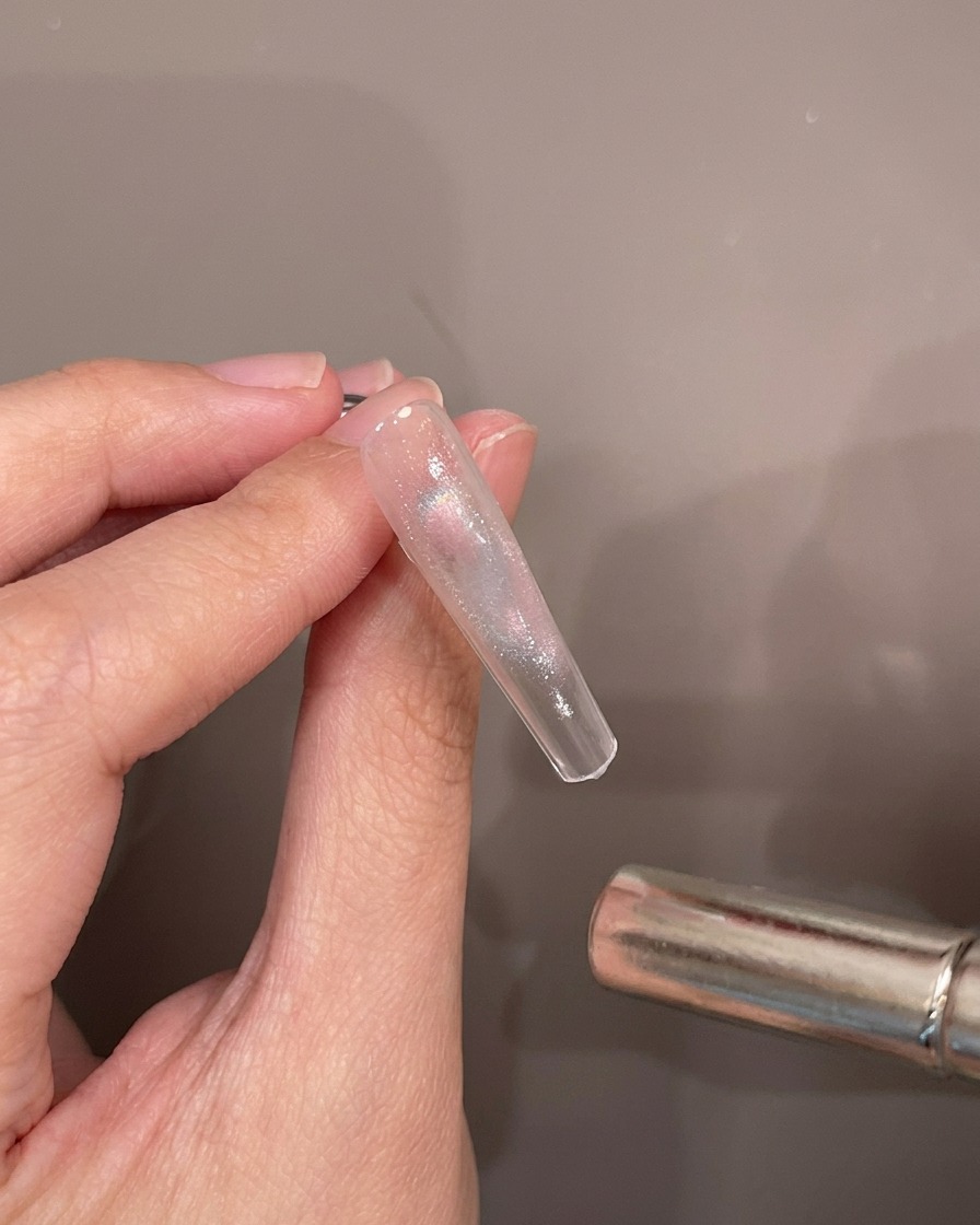 Eine Hand hält einen klaren Nageltip, während ein Magnetstab über das nasse Gel schwebt.