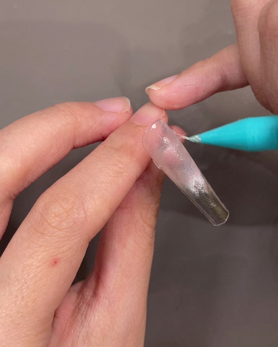 Nahaufnahme einer Hand, die eine dünne, diagonale Linie aus silbernem Glitzergel über einen klaren Nageltip zieht.
