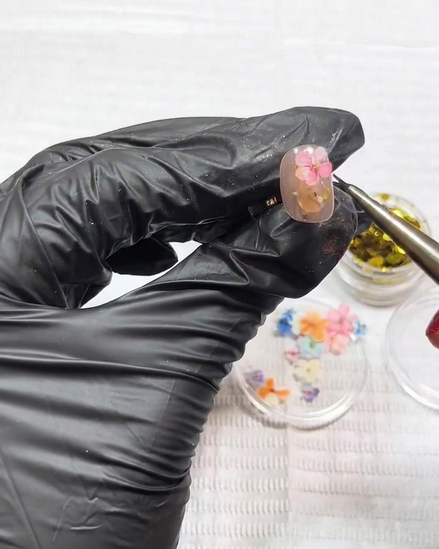 Eine Pinzette setzt eine winzige, flache rosa Trockenblume vorsichtig auf das feuchte Gel auf.