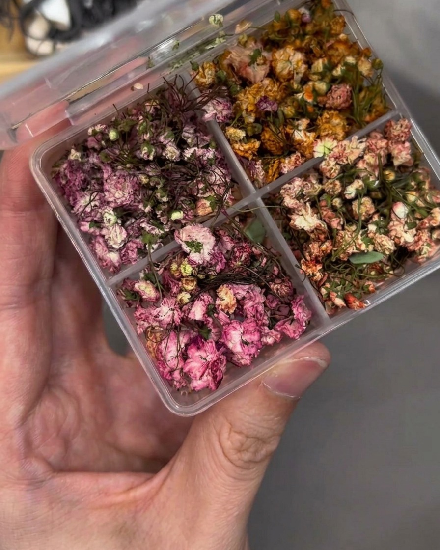 Eine Pinzette platziert vorsichtig eine kleine Trockenblume auf einem Nagel-Tip.