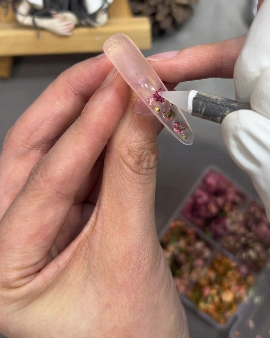 Nahaufnahme einer Hand, die kleine Goldperlen und rosafarbene Strasssteine auf einen klar beschichteten Nagel-Tip setzt.