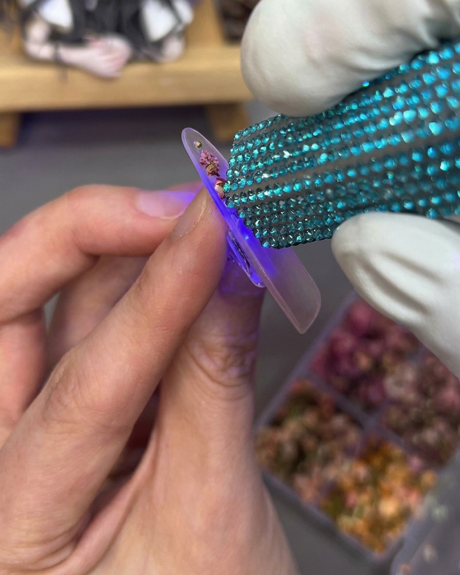 Eine Hand hält eine Mini-UV-Lampe über einen Nagel-Tip, auf dem Blumen und Strasssteine eingebettet sind.
