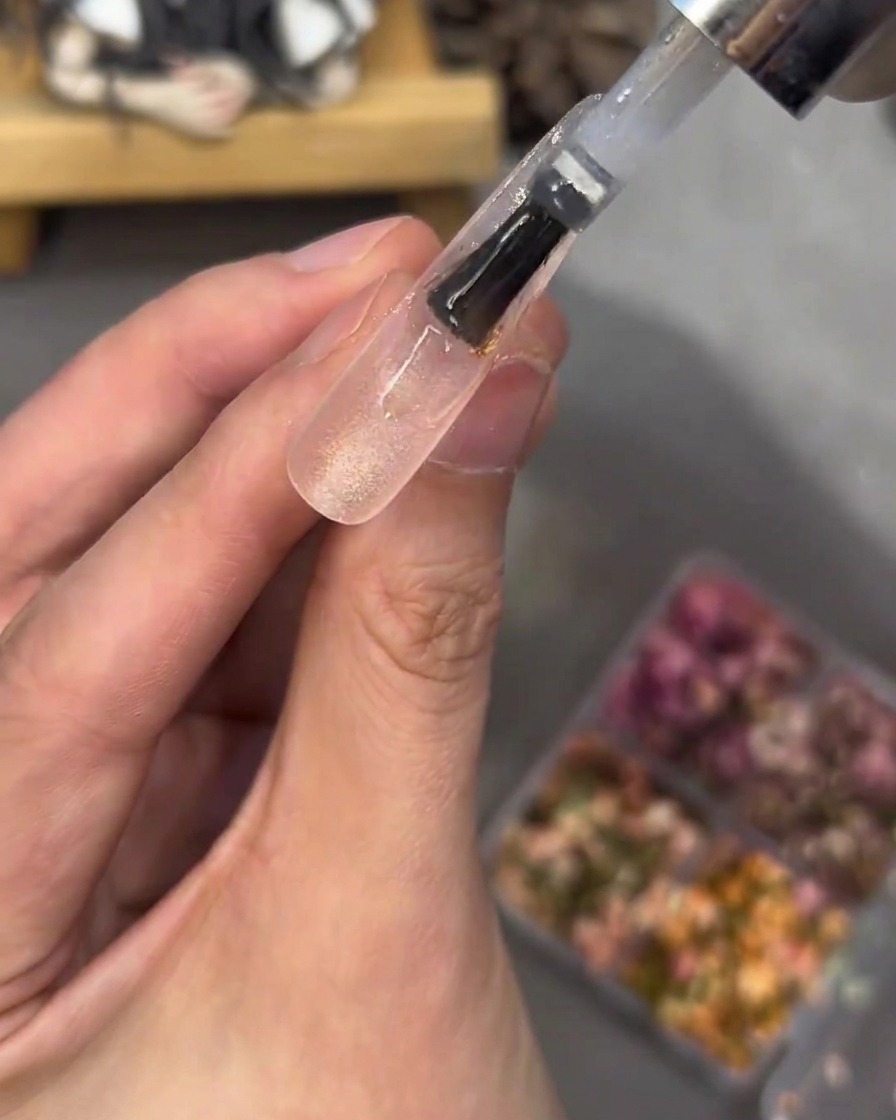 Ein Pinsel trägt eine klare, dicke Gelschicht über den mit Blumen und Strasssteinen verzierten Nagel-Tip auf.