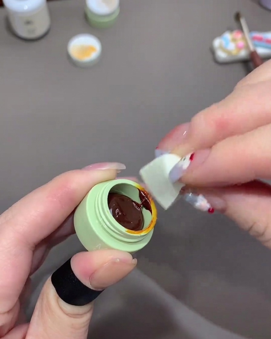 Eine Hand benutzt ein kleines Stück eines weißen Make-up-Schwämmchens, um dunkelorangefarbenes Gel aus einem kleinen grünen Tiegel aufzunehmen.