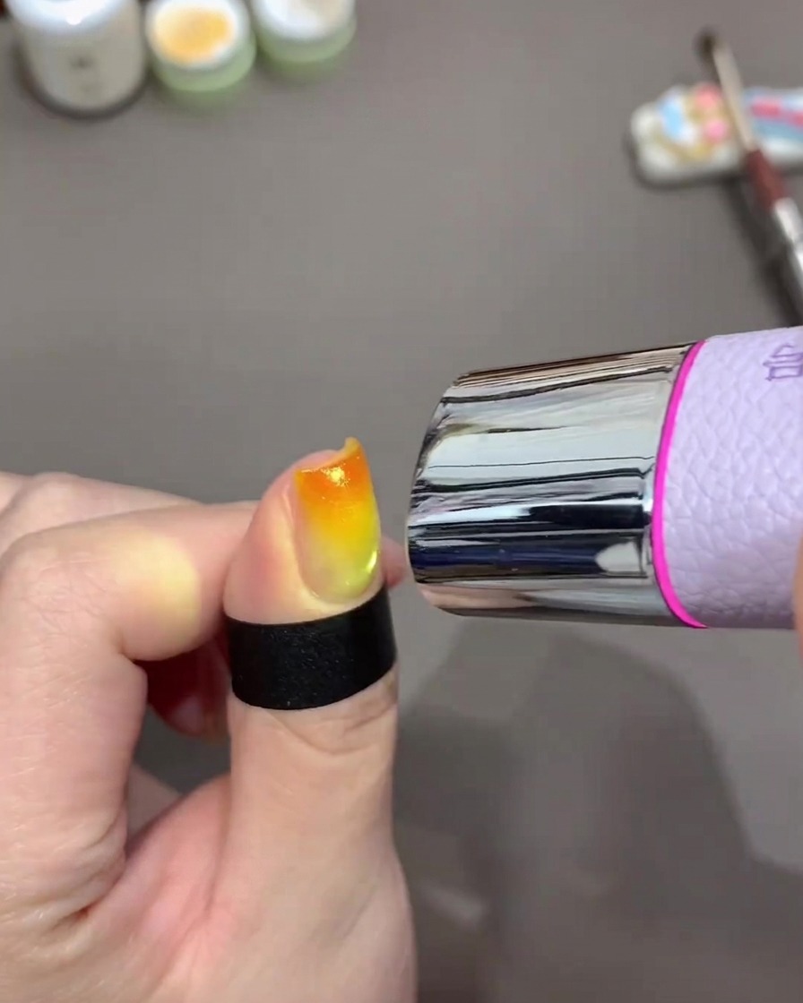 Eine kleine Hand-UV-Lampe ist über einem Fingernagel positioniert, um ein orange-gelbes Gradient-Design auszuhärten.