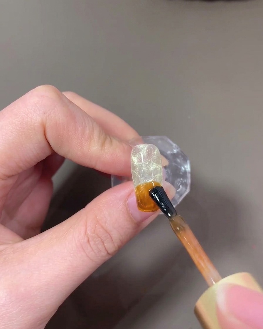 Ein Pinsel trägt bernsteinfarbenes, transluzentes Gel auf die Nagelspitze auf.