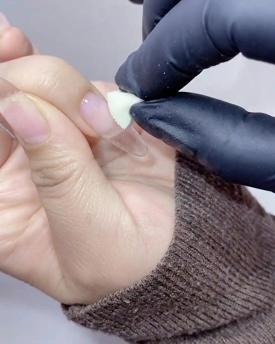 Behandschuhte Hände verwenden ein kleines Kosmetikschwämmchen, um pfirsich-oranges Gel auf die Mitte eines transparenten Nagels zu tupfen.