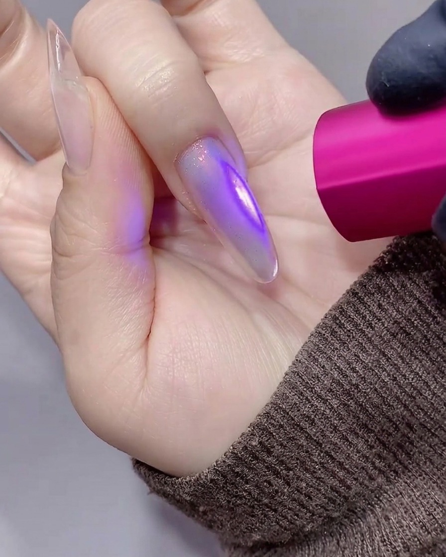 Nahaufnahme einer Hand mit einem langen, violett-getönten Stiletto-Nagel, der unter einer kleinen pinken UV/LED-Handlampe ausgehärtet wird.