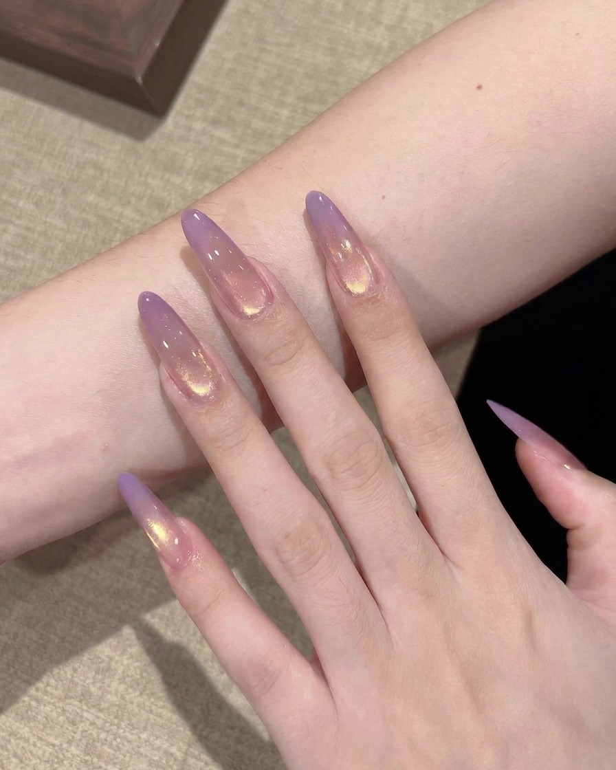 Nahaufnahme eines flachen Pinsels, der einen kleinen Tropfen transparentes, lilafarbenes Gel auf die Spitze eines klaren Stiletto-Nagel-Extensions aufträgt.