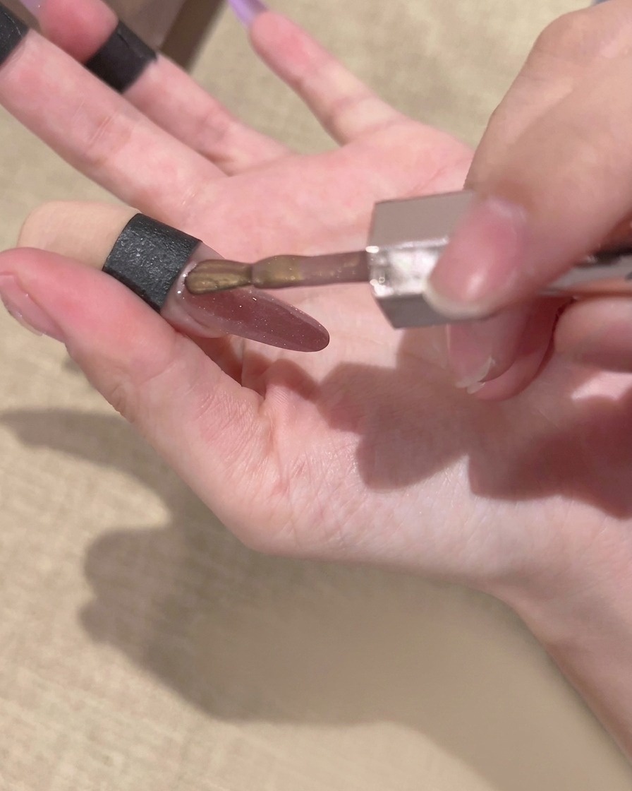 Ein Pinsel aus einer Gelpoliturflasche trägt eine Schicht goldenes, magnetisches Cat-Eye-Gel über einem Nagel mit bestehendem lila Farbverlauf auf.