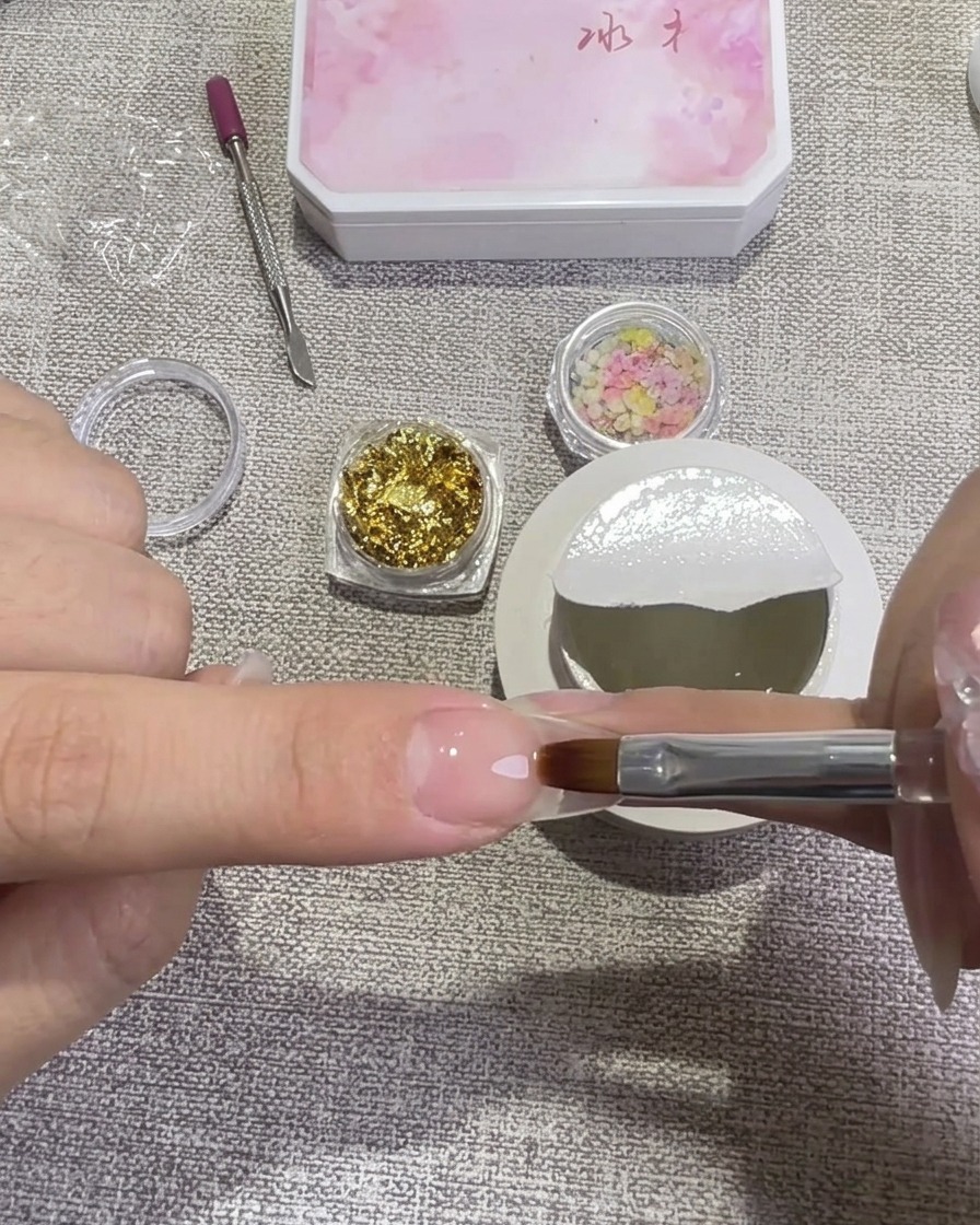 Eine Hand verwendet einen flachen Gelpinsel, um eine Schicht klares Hard Gel auf einen vorbereiteten Nagel aufzutragen, mit kleinen Döschen voller Blattgold und Trockenblumen im Hintergrund.