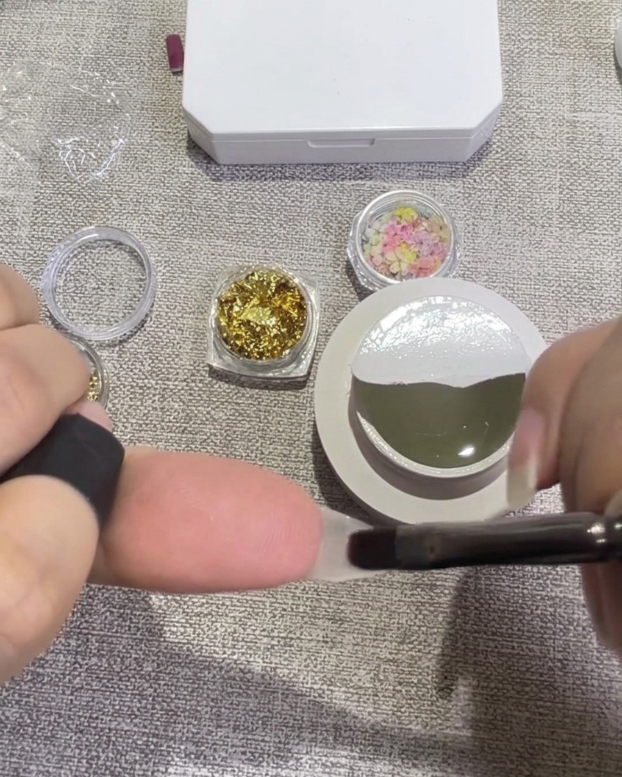 Eine Pinzette platziert kleine, bunte Trockenblumenblätter auf einem mit feuchtem, klarem Gel bedeckten Nagel.