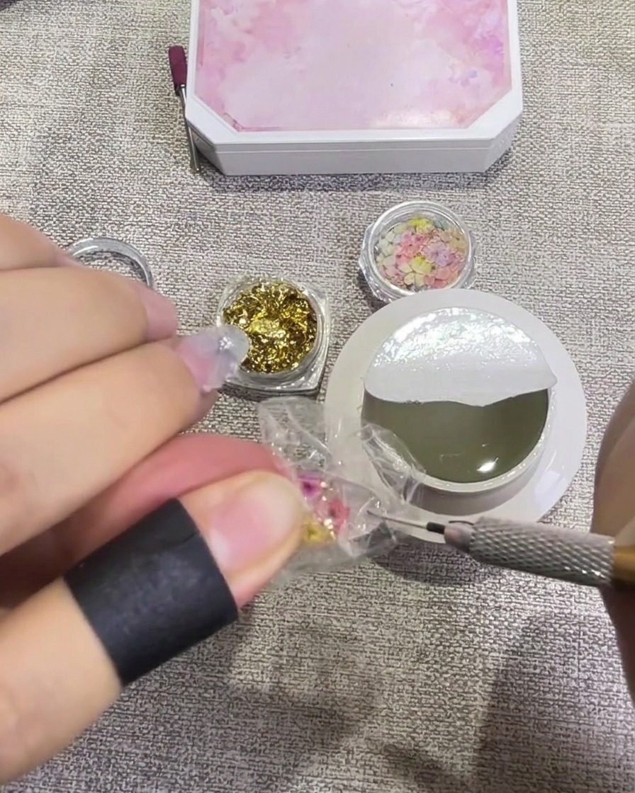 Ein Pinsel trägt einen glänzenden, klaren Top Coat über einen mit gepressten Blumen und Blattgold verzierten Nagel auf, wodurch er nass und transparent aussieht.