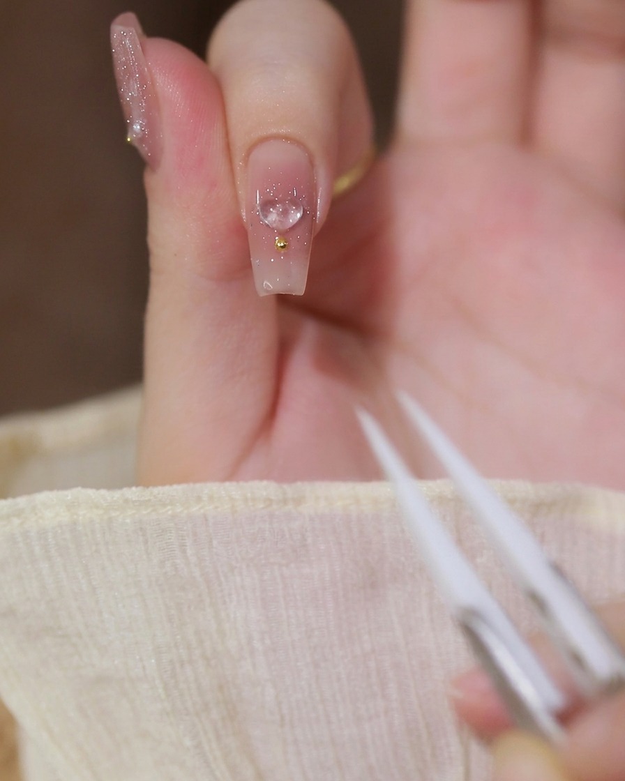 Nahaufnahme eines Zeigefingers mit milchig-nude Gel-Basis, einem transparenten 3D-Herz-Charm und einer Goldperle.