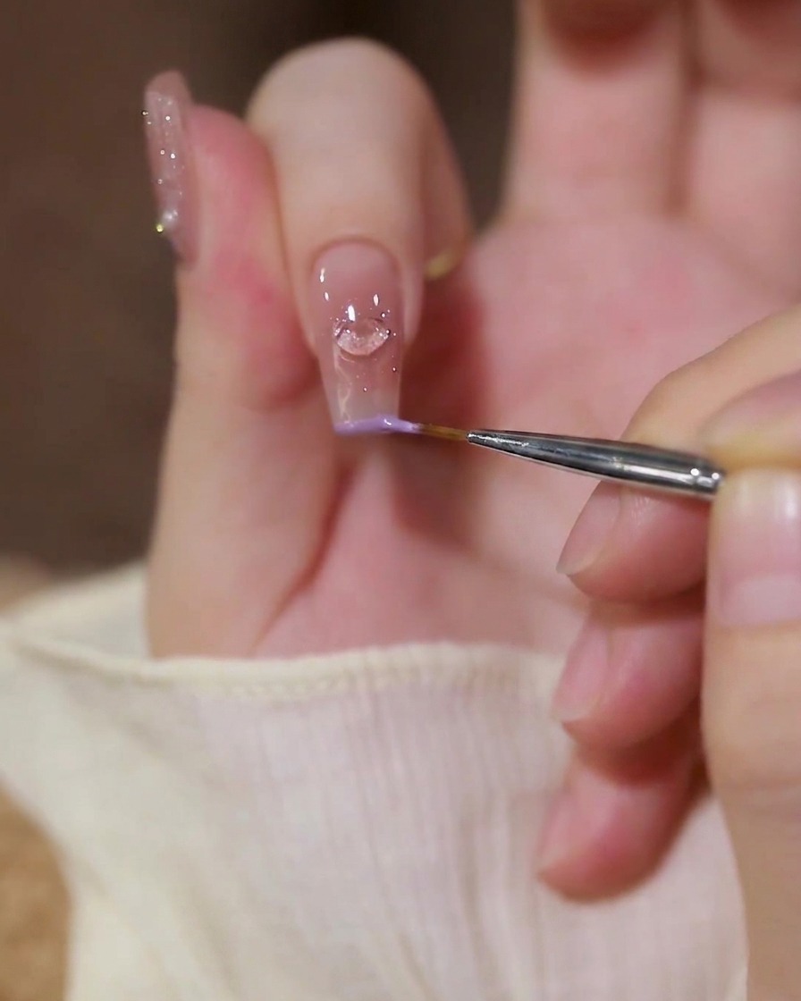 Nahaufnahme eines feinen Liner-Pinsels, der pastell-lila Gel-Nagellack entlang der Kante eines Zeigefingers mit Herz-Charm aufträgt.