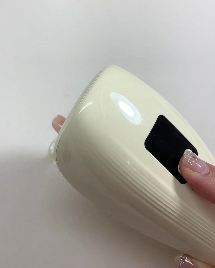 Eine Hand hält eine kleine, weiße UV/LED-Lampe direkt über einen frisch lackierten Nude-Nagel.