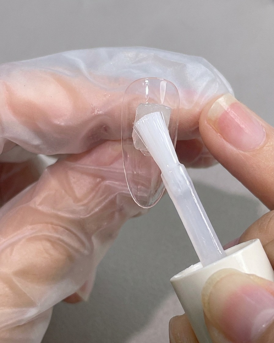 Ein Pinsel trägt klares, viskoses Blooming Gel diagonal über eine Nagelspitze auf.