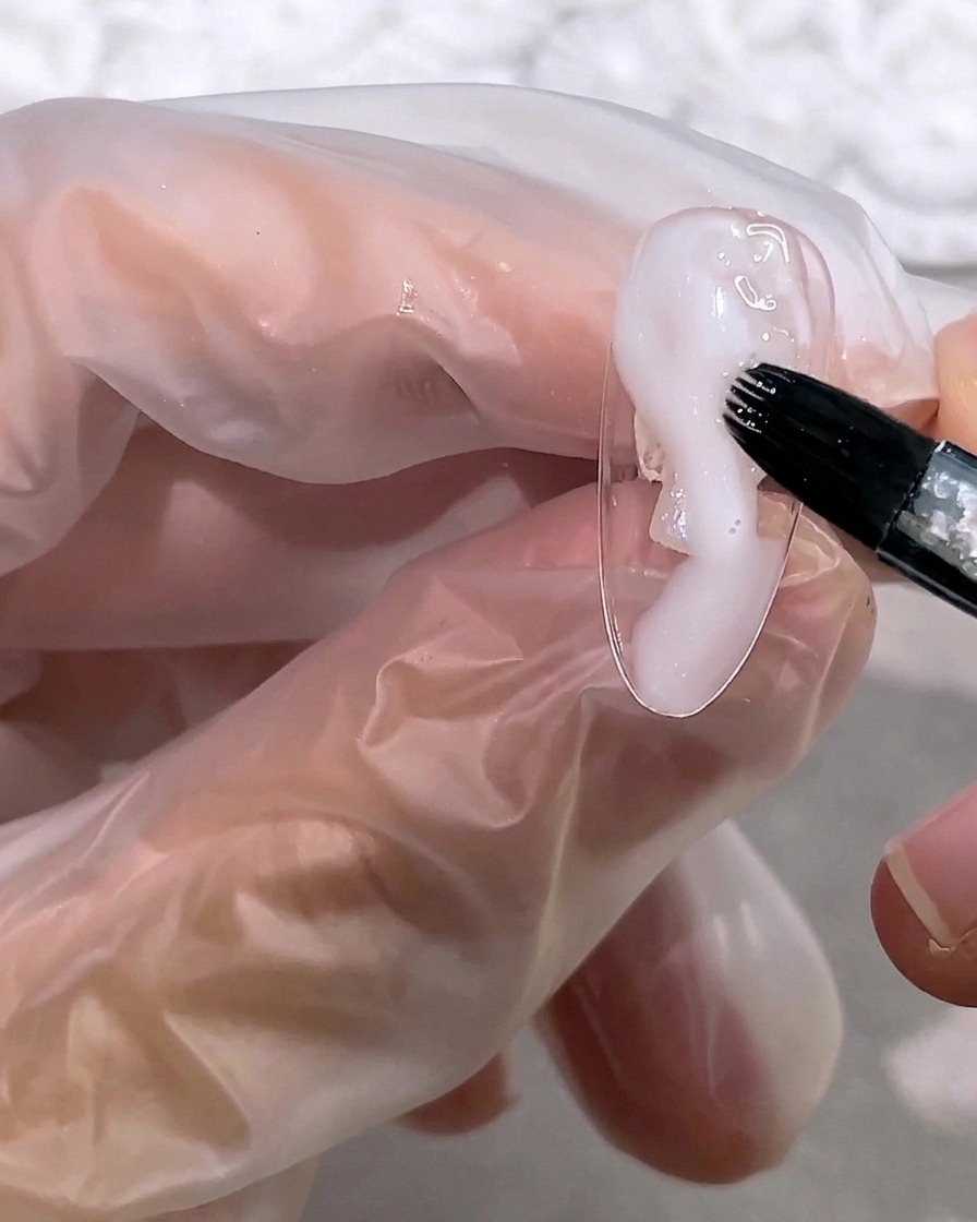 Ein Pinsel trägt eine kleine Menge milchiges, halbtransparentes weißes Gel auf die Nagelspitze auf.