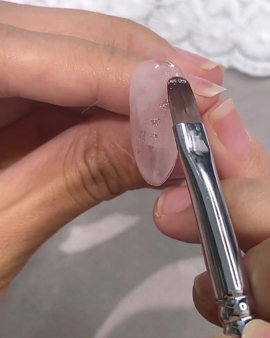 Ein Pinsel trägt ein transparentes rosa Gel über einen klaren Nagel mit Silberflocken auf.