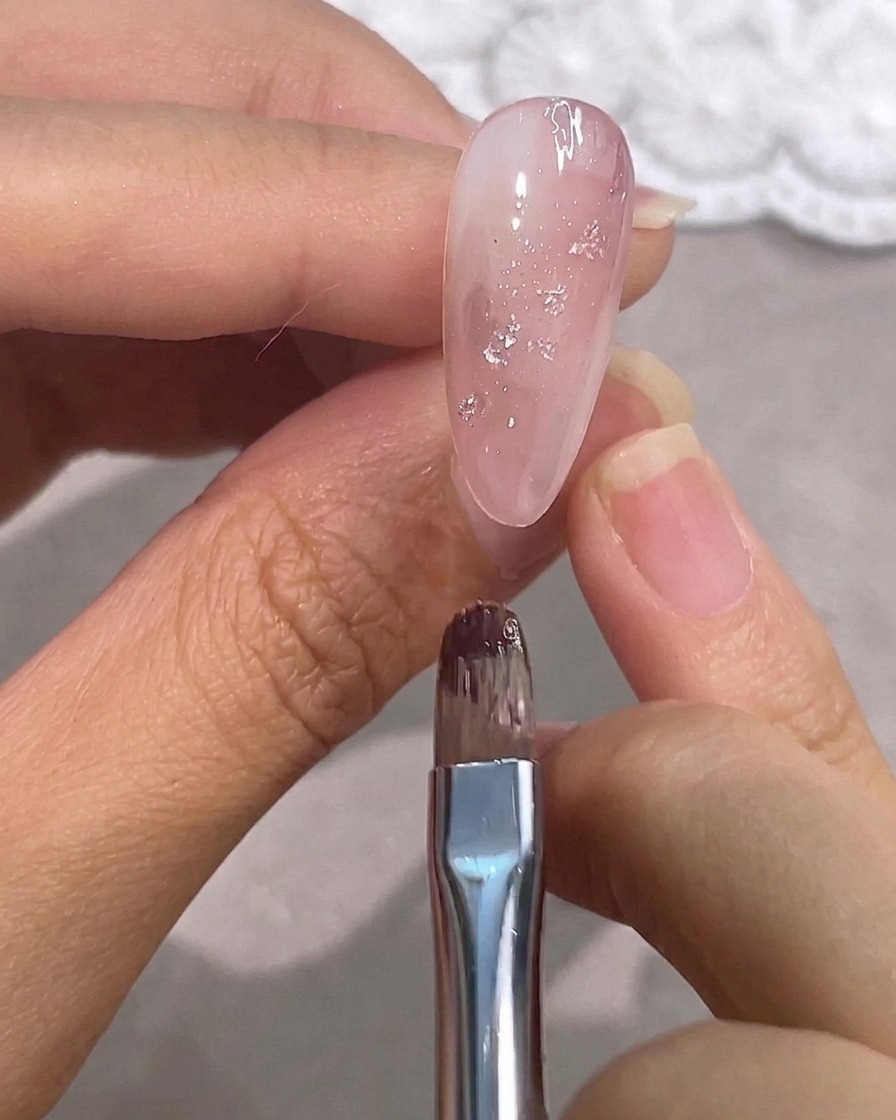 Ein Detailpinsel über einem glänzenden rosa Gelnagel mit Silberfolien-Akzenten.