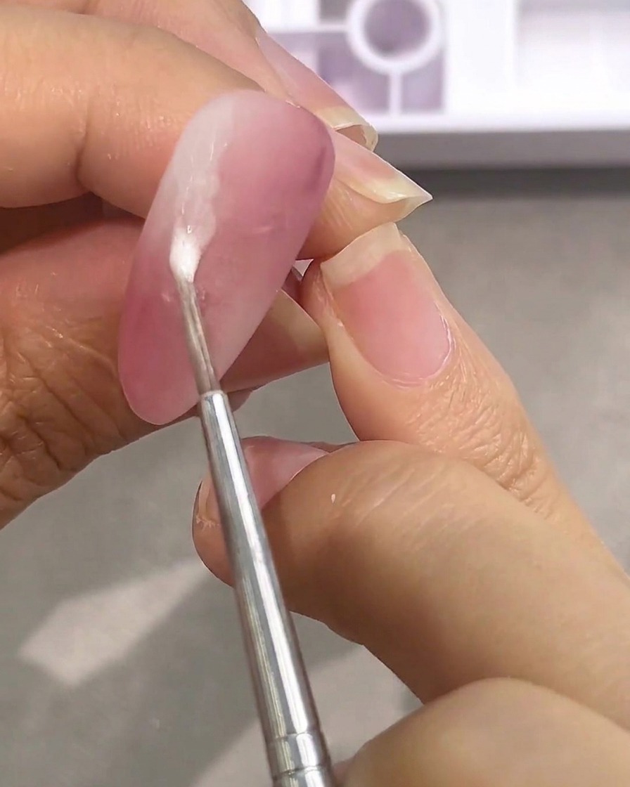 Ein Werkzeug trägt einen Tropfen wässrige weiße Farbe auf einen matten rosa Nagel auf.