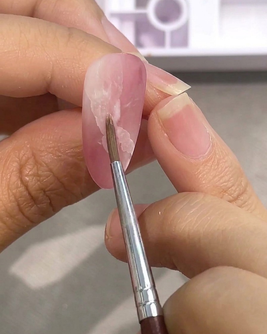 Ein feiner Detailpinsel verblendet die Kanten eines weißen Aquarell-Flecks auf einem matten rosa Nagel mit Wasser.