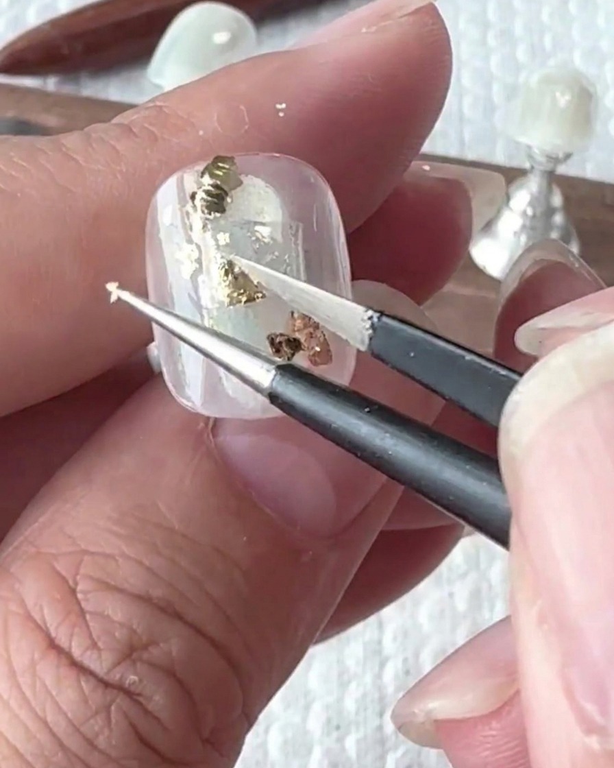 Eine Pinzette platziert vorsichtig unregelmäßige Stücke Goldfolie auf der Nageloberfläche.