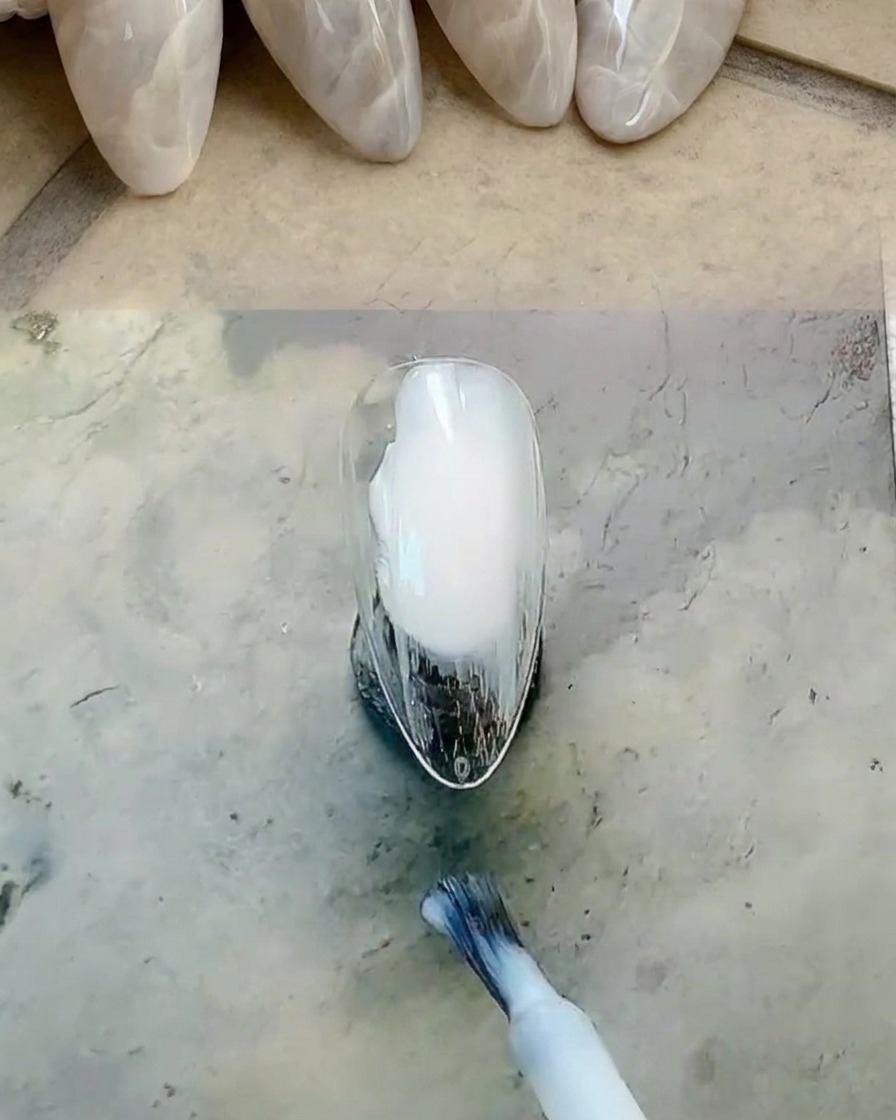 Ein Flaschenpinsel trägt milchig-weißen Gel-Lack auf die Nageloberfläche auf.