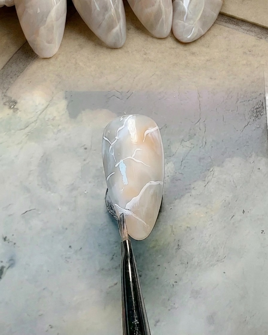 Ein kleiner, sauberer Rundpinsel verwischt die Kante einer weißen Gel-Linie auf einem marmorierten Nagel.