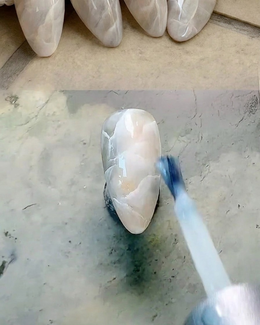 Ein flacher Pinsel trägt eine dünne Schicht milchig-weißes Gel über einem marmorierten Nagel auf.