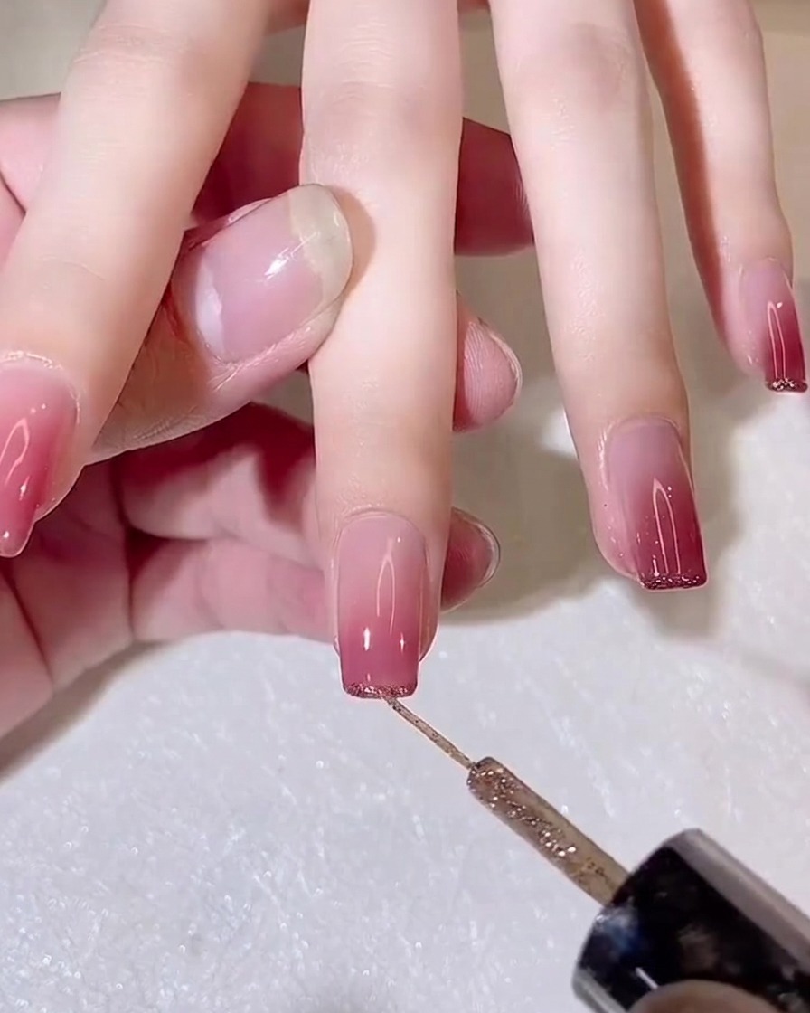 Ein feiner Liner-Pinsel trägt eine dünne Linie Goldglitzer-Gel auf die freie Kante eines Gradient-Nagels auf.