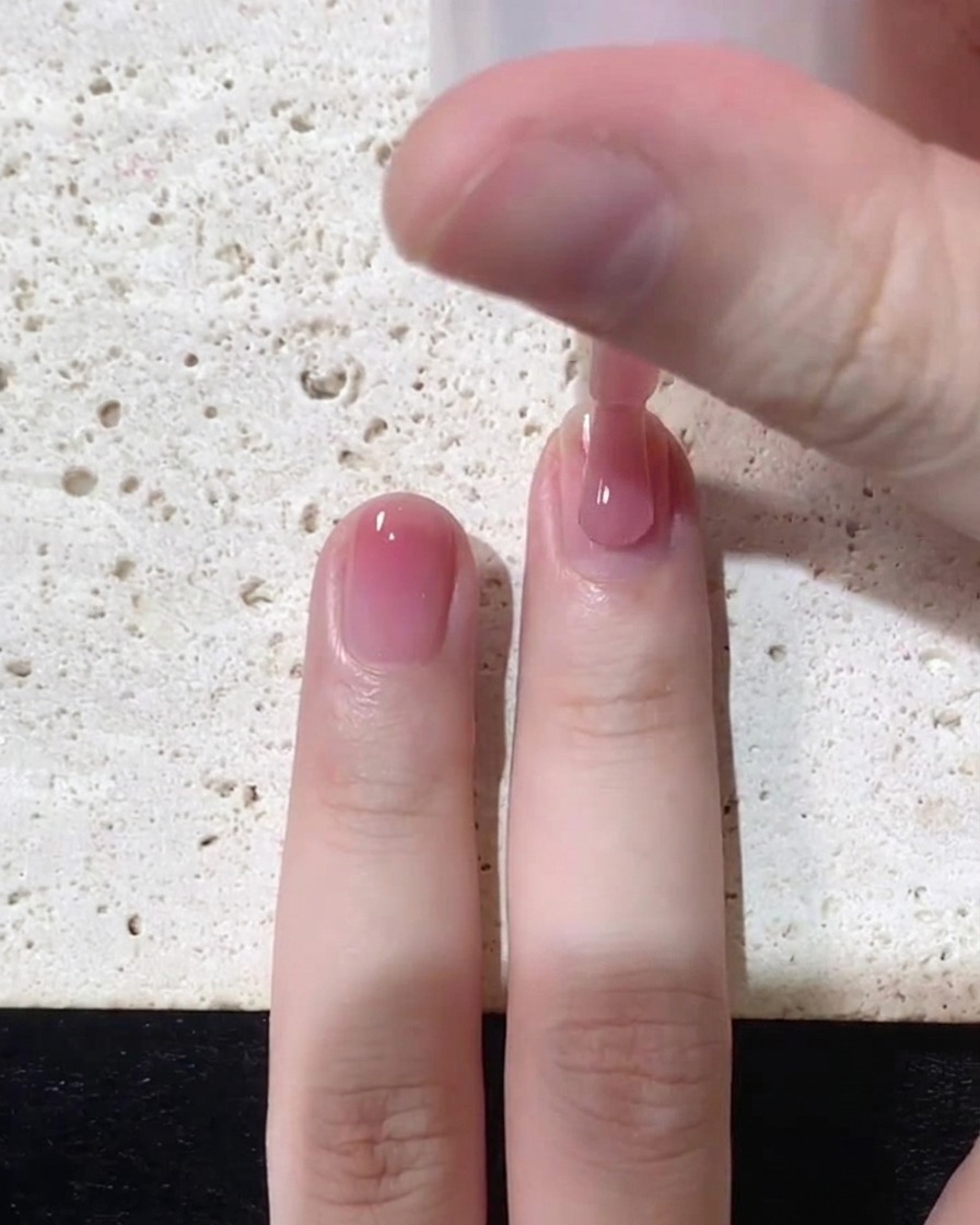 Nahaufnahme eines Pinsels, der transparenten rosa Jelly-Lack nahe am Nagelbett auf einen Naturnagel aufträgt.