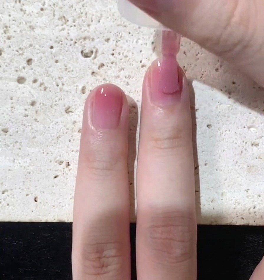 Nahaufnahme, wie ein Pinsel eine dünne Schicht durchscheinenden rosa Lacks über den gesamten Naturnagel aufträgt.