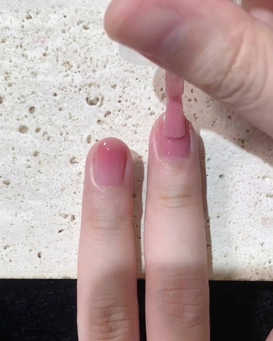 Ein Pinsel streicht den rosa Jelly-Lack gleichmäßig zur Nagelspitze über die obere Hälfte des Nagels.