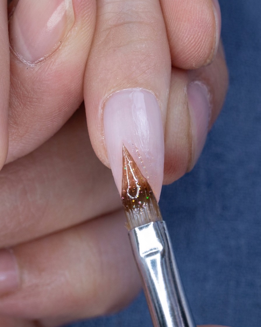 Nahaufnahme eines feinen Pinsels, der kleine Glitzerpartikel auf die Spitze eines Nude-Rosa-Ombre-Nagels aufträgt.