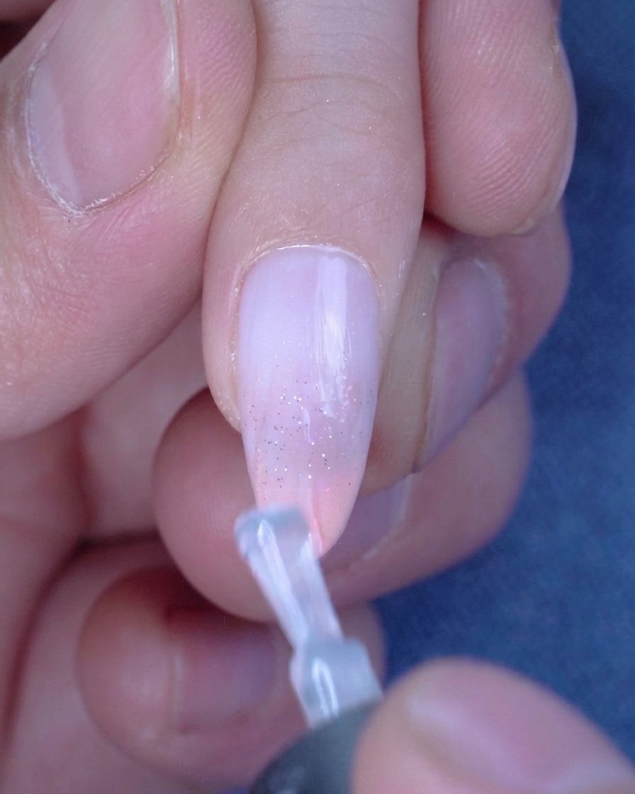 Ein Pinsel trägt eine glatte, klare Schicht Top Coat auf einen mit Glitzer verzierten Ombre-Nagel auf.