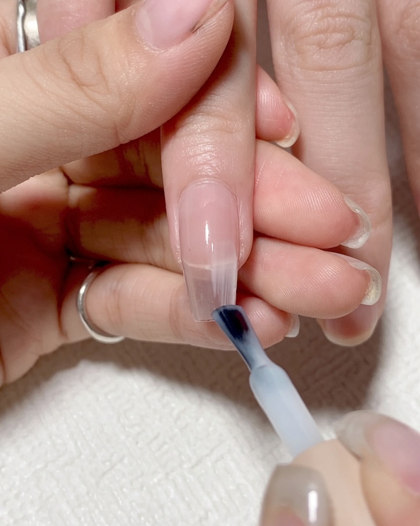 Ein Nagelpinsel trägt ein durchscheinendes, helles Gel auf die rechte Seite des Nagels auf.
