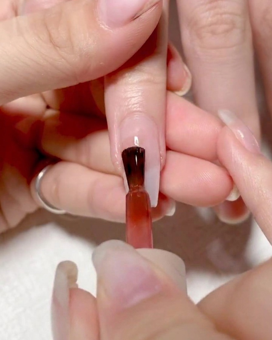 Ein Nagelpinsel trägt leuchtend rotes Gel auf die linke Seite des Nagels auf.