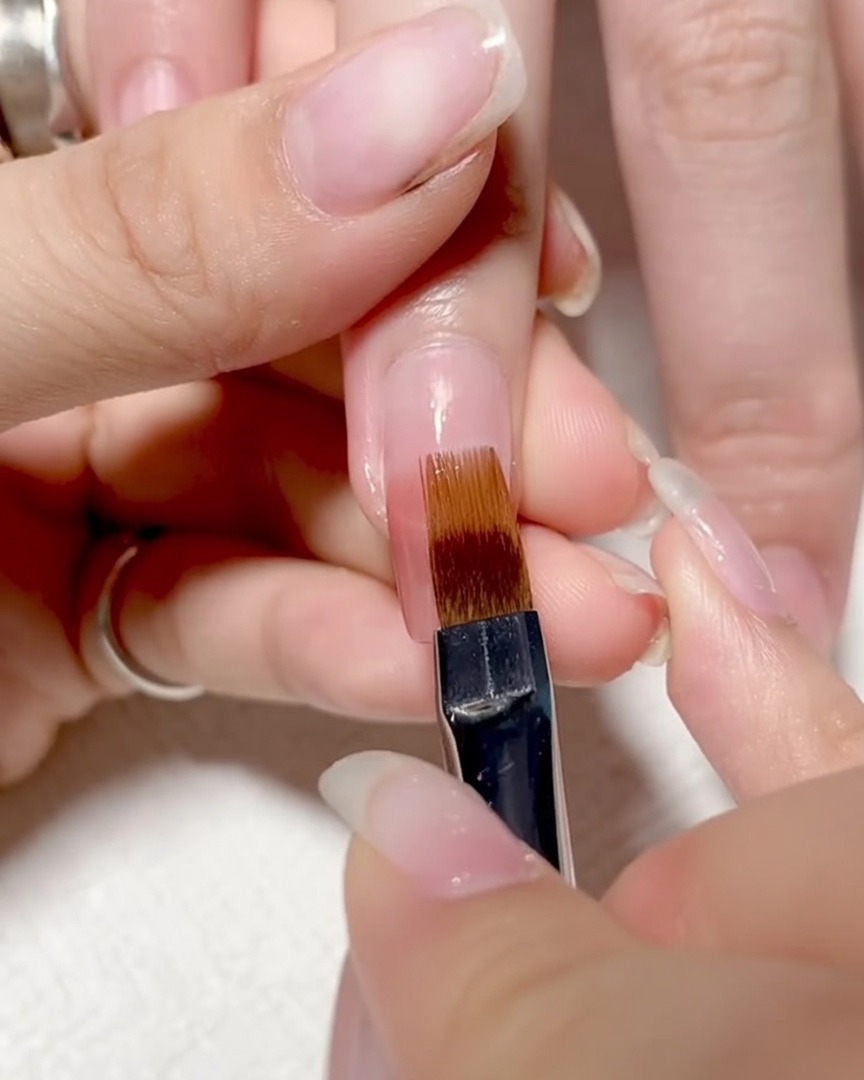 Ein Pinsel wird sanft über die Nageloberfläche nach unten gezogen, um die Farben zu einem glatten Ombré zu verblenden.