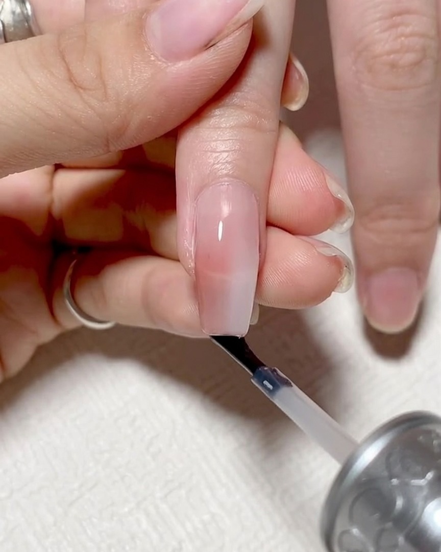 Eine Nahaufnahme zeigt, wie der Nagelpinsel horizontal an der freien Nagelkante entlangfährt, um diese zu ummanteln.