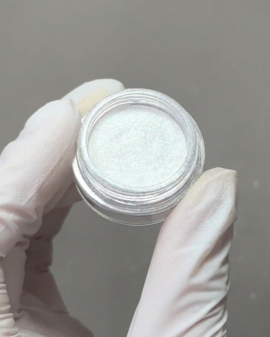 Eine behandschuhte Hand hält einen kleinen Behälter mit irisierendem Aurora-Chrom-Puder, während ein Schwamm-Applikator etwas Pigment aufnimmt.